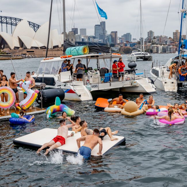 A group of people enjoying a lively Yacht Social Club event in a harbor with the Sydney Opera House and Sydney Harbour Bridge in the background. They are swimming, lounging on floating mats, and using colorful inflatables. The scene is festive and vibrant.