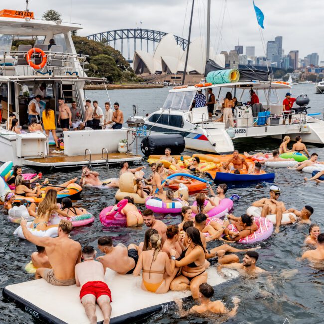 A lively scene of people enjoying a boat party on a river with Sydney Opera House and Harbour Bridge in the background. The revelers are swimming, lounging on inflatables, and socializing on a floating platform and near several boats. The sky is overcast, showcasing the vibrant atmosphere of The Yacht Social Club Sydney Boat Hire.