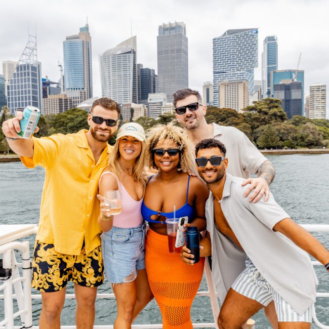 Five people are standing on a boat with a city skyline in the background. They are smiling, holding drinks, and wearing casual summer clothing. The sky is partly cloudy. The group appears cheerful and celebratory, enjoying a day out on the water with The Yacht Social Club Sydney Boat Hire.