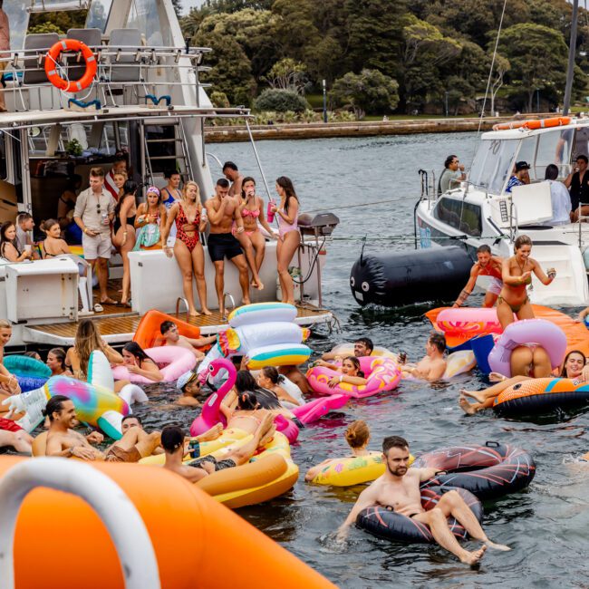 A lively scene shows people on inflatable floats of various shapes and colors in the water, surrounded by several boats. Some participants are on the boats, while others are enjoying the water, suggesting a festive outdoor gathering reminiscent of The Yacht Social Club Event Boat Charters.