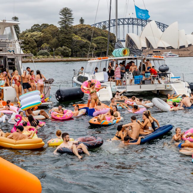 A vibrant scene of a floating party with numerous people on inflatable rafts and toys in the water. They are gathered between several boats from The Yacht Social Club, with the Sydney Opera House and Harbour Bridge visible in the background on a sunny day.