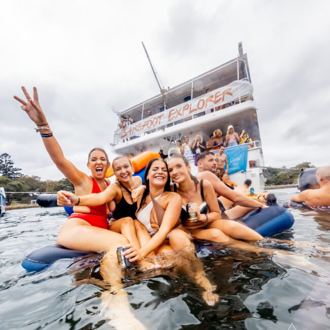 Four women smiling and posing while sitting on an inflatable raft in the water, with one holding up a peace sign. Behind them, people are gathered on a large boat named "Barefoot Explorer." The background shows a cloudy sky and tree-covered shoreline. Text: "The Yacht Social Club Event Boat Charters.