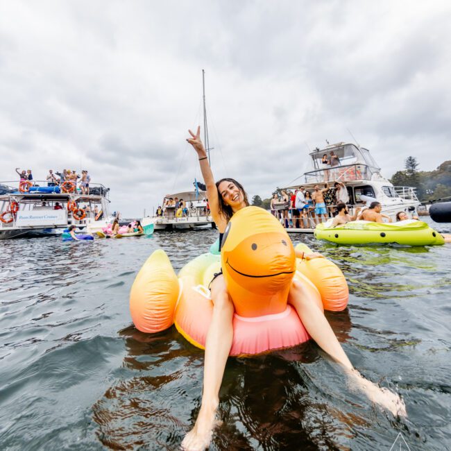 A person is sitting on an inflatable duck float in the water, smiling and making a peace sign with their hand. In the background, groups of people are on yachts and other inflatables from The Yacht Social Club Event Boat Charters, with a cloudy sky overhead. The setting is festive and lively.