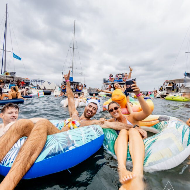 A group of people float on inflatable rafts in the ocean near several yachts under a cloudy sky. Two smiling individuals in the foreground are taking a selfie; one wears sunglasses, and the other sports a tropical shirt. The scene exudes a lively, fun atmosphere, much like Boat Parties Sydney The Yacht Social Club.