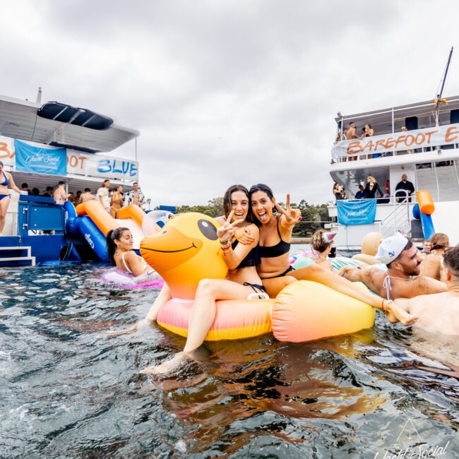 Two women smile and flash peace signs while sitting on an inflatable golden duck in the water. Several people float and swim around them. A boat party with the banner "BAREFOOT & BLUE" can be seen in the background, hosted by The Yacht Social Club.