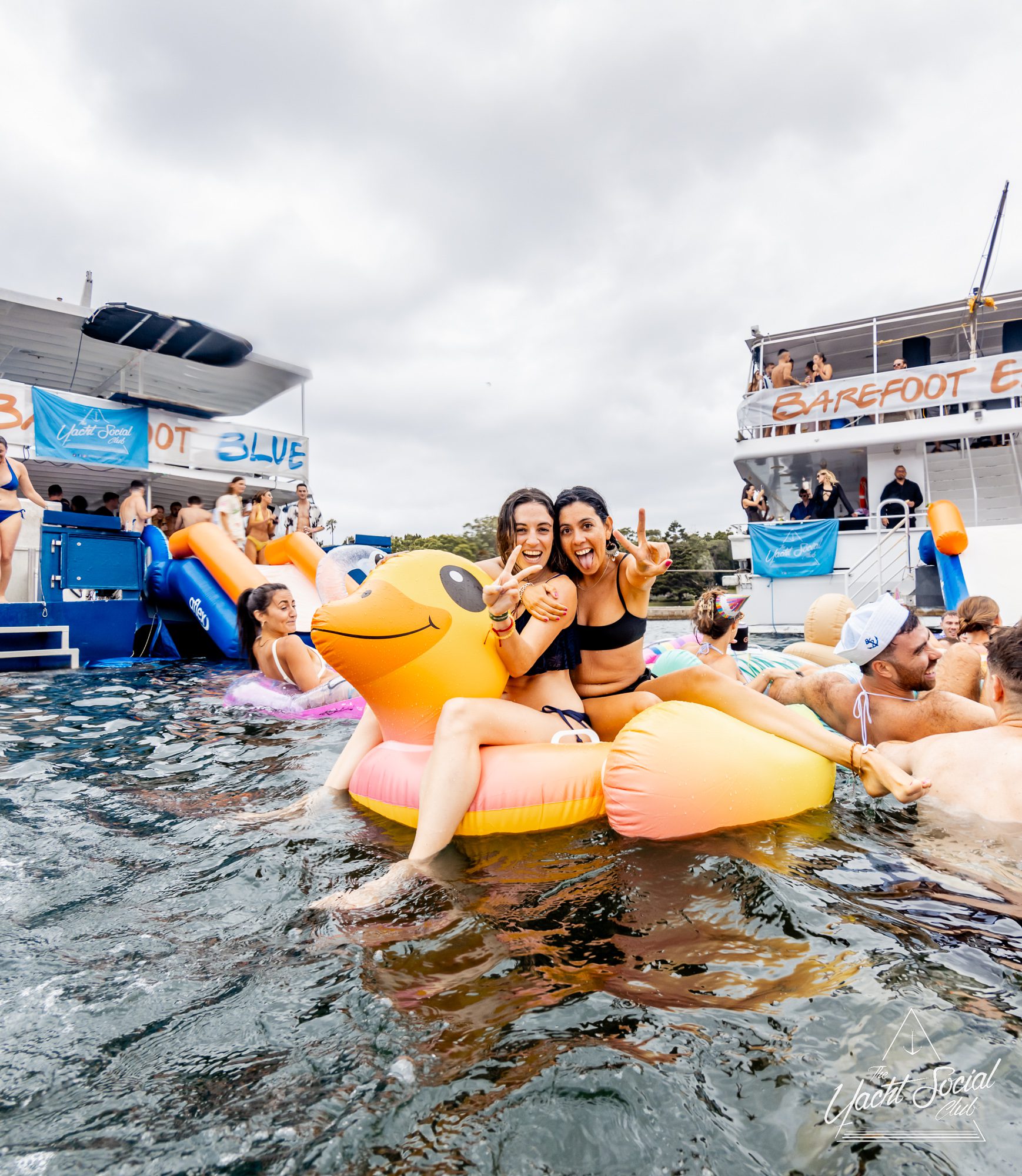 Two women smile and flash peace signs while sitting on an inflatable golden duck in the water. Several people float and swim around them. A boat party with the banner "BAREFOOT & BLUE" can be seen in the background, hosted by The Yacht Social Club.