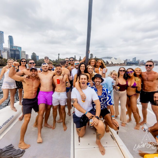 A group of smiling people in casual summer attire, including swimsuits and shorts, are gathered on a boat with a city skyline and a bridge in the background. They are enjoying drinks and appear to be celebrating with The Yacht Social Club. The sky is cloudy.