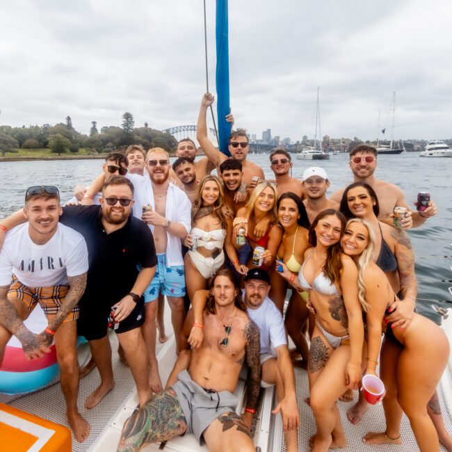 A group of people are gathered on a boat, smiling and posing for the camera. The background features a waterfront with trees and a cloudy sky. Some individuals are holding drinks while others are dressed in swimsuits and summer attire, enjoying a festive atmosphere at Boat Parties Sydney The Yacht Social Club.