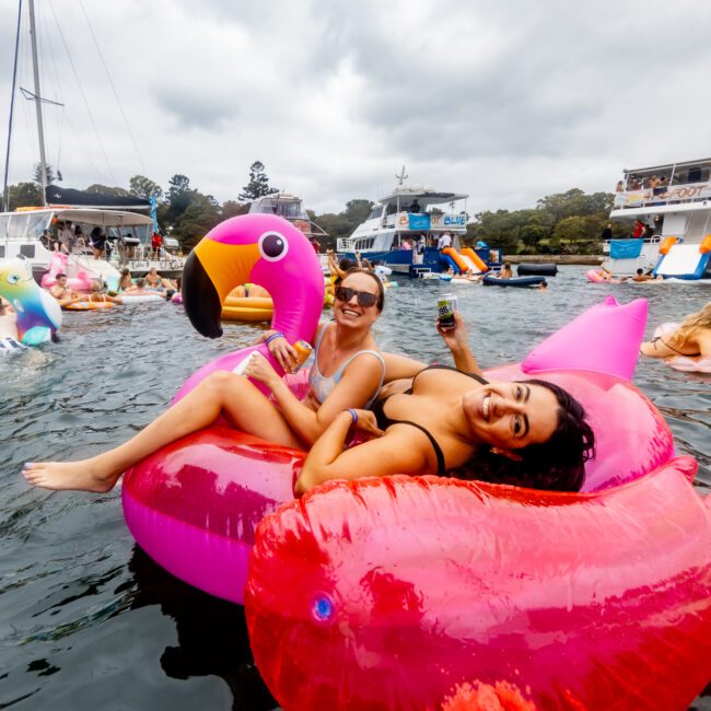 Two women are lounging on a large pink flamingo float in a lively water setting, surrounded by others on floats and boats, enjoying The Yacht Social Club Event Boat Charters. The sky is overcast, adding a contrasting backdrop to the colorful scene.