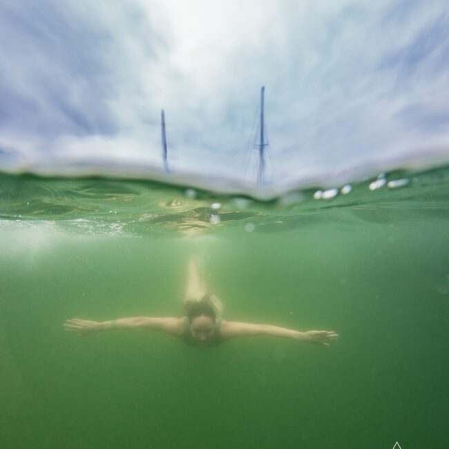 A swimmer is underwater with outstretched arms in greenish water, the surface breaking to reveal a cloudy sky and sailboat masts above. The logo of "The Yacht Social Club Sydney Boat Hire" is visible in the bottom right corner.