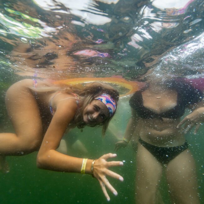 Underwater photo of two people in swimsuits. The person on the left is reaching towards the camera with a smiling expression and a colorful headband. The person on the right is partially visible, wearing a black swimsuit. The clear water and sunlight filtering through hint at an ideal day for Sydney Harbour Boat Hire the Yacht Social Club experience.