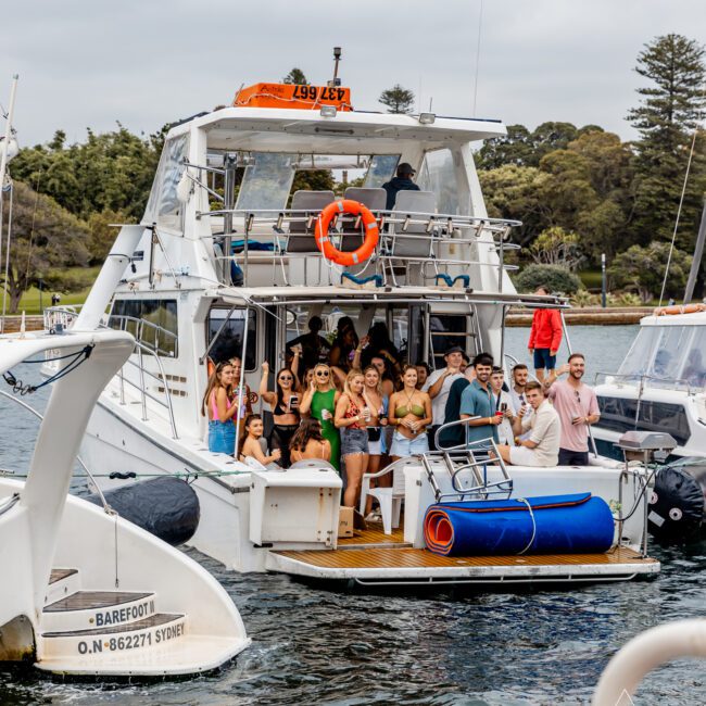 A group of people is gathered on the deck of a white yacht named "Barefoot." The yacht is docked in a marina, surrounded by water and other boats. Everyone appears to be enjoying a party, with some holding drinks. Trees and greenery are visible in the background, epitomizing Luxury Yacht Rentals Sydney.