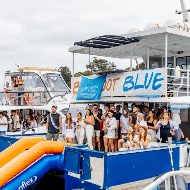 A lively boat party by "BLUE" features guests enjoying an orange and blue inflatable slide. Nearby boats and a coastal backdrop complete the scene, with participants in summer attire smiling and mingling. Experience Boat Parties Sydney with The Yacht Social Club for unforgettable memories!