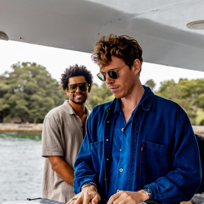 Two men on a boat, with one in sunglasses and a blue shirt focused on DJ equipment, while the other, also in sunglasses and a beige shirt, smiles in the background. Water and trees are visible in the distance. Experience moments like these with The Yacht Social Club Event Boat Charters.