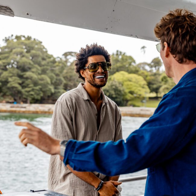 Two men are having a lively conversation on the deck of The Yacht Social Club boat. One man, with sunglasses and a light-colored shirt, is smiling while standing near the railing. The other man, also wearing sunglasses and a blue jacket, faces him and gestures with one hand. DJ equipment is visible on a table nearby.