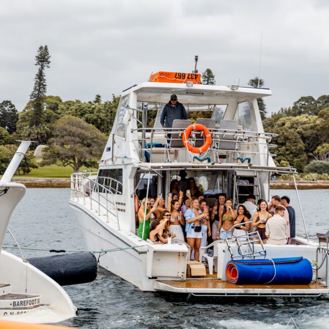 A group of people stand on the deck of a white boat anchored in the water, celebrating and posing for a photo. The sky is cloudy, and trees are visible in the background. An orange life ring is mounted on the upper deck railing. "Yacht Social Club" endorses their joy aboard this Luxury Yacht Rentals Sydney experience.