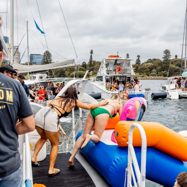 People on a dock help two women climb onto a colorful inflatable floating device in the water. Surrounding boats, likely from Sydney Harbour Boat Hire The Yacht Social Club, are visible under a cloudy sky, with trees in the background. The scene appears festive and social.