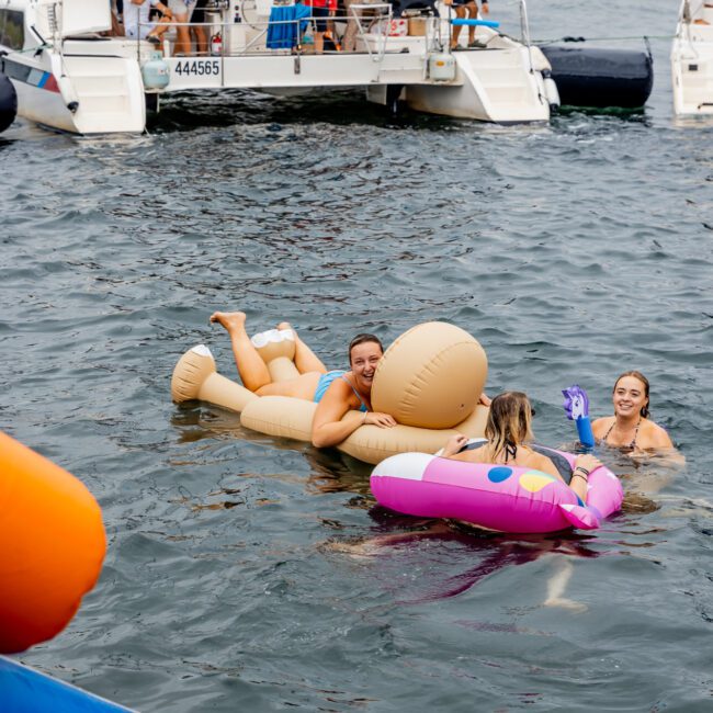 People are enjoying a sunny day on the water, lounging on inflatable pool toys with yachts anchored nearby. The iconic Sydney Opera House is visible in the background, part of a vibrant Yacht Social Club event. Experience the ultimate fun with Sydney Harbour Boat Hire by The Yacht Social Club.