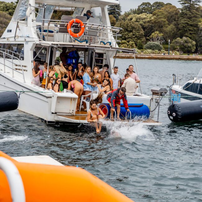 A large group of people are enjoying a lively party on a white yacht. Some are on the deck, others are entering the water, surrounded by greenery and a calm body of water. An orange and blue inflatable object is visible in the foreground, adding to the fun vibe at The Yacht Social Club Sydney Boat Hire event.