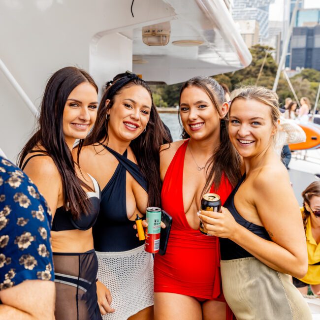 Four smiling women in swimwear pose together on a boat. The first woman wears a black bikini top with a sheer cover-up skirt, the second woman wears a black bikini, the third woman wears a red one-piece swimsuit, and the fourth woman wears a beige bikini. Experience fun like this with The Yacht Social Club Sydney Boat Hire!