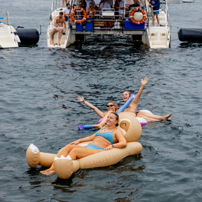 Three people are floating on large, inflatable pool loungers in a body of water, joyfully raising their arms. Behind them, a group of people are gathered on a boat enjoying the day. The background shows city buildings and other boats. Text on the image reads "The Yacht Social Club Sydney Boat Hire".