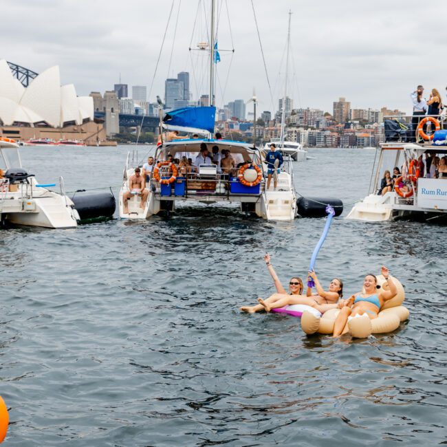 People floating on inflatable rings in the water near anchored boats. In the background, the Sydney Opera House and Harbour Bridge are visible against the city skyline. The scene is lively with swimmers and people on the boats enjoying boat parties hosted by The Yacht Social Club.