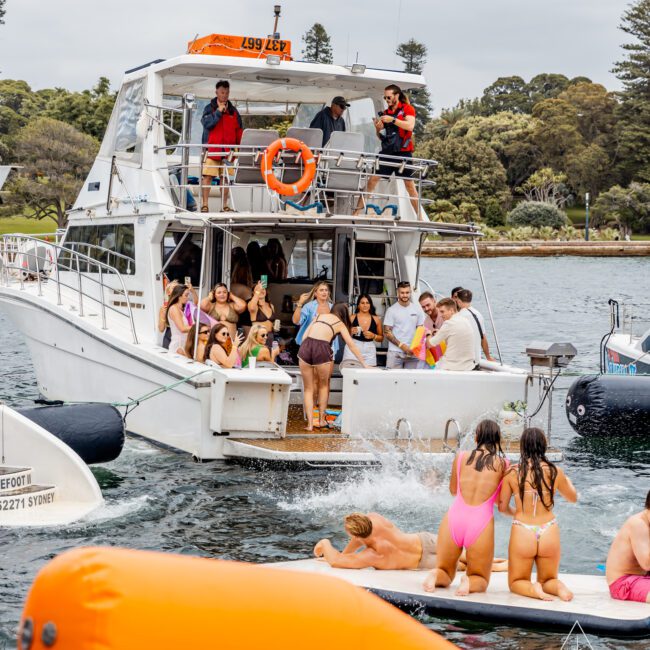 A lively group enjoys a boat party hosted by The Yacht Social Club in Sydney, with some participants diving and swimming. The boat is docked in a scenic, tree-surrounded area, and the atmosphere is cheerful with everyone in swimsuits enjoying their time.