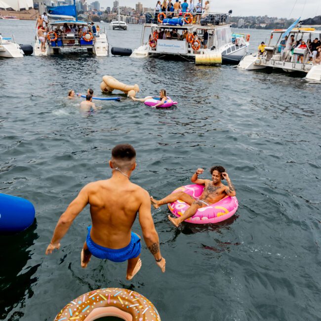 People enjoy a party on anchored yachts with inflatables in the water. A man jumps into the water near a donut-shaped float, while others relax on an inflatable pink flamingo. The background includes more boats and the Sydney Opera House, all part of The Yacht Social Club Sydney Boat Hire.