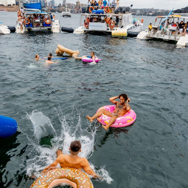 A lively scene on the water shows several anchored yachts from The Yacht Social Club filled with people partying. In the foreground, individuals float on inflatable rings, including a donut and a flamingo, while others swim and splash in the water. The skyline is visible in the background.