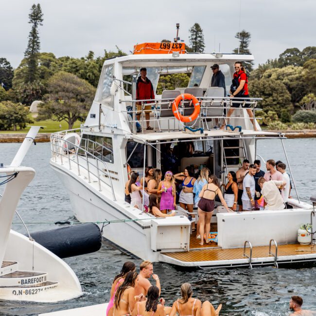 A group of people is enjoying a day on a white yacht named "Barefoot," anchored near the shore. Some individuals are on the lower deck, while others are dancing or lounging on a floating platform in the water. Trees and a park are visible in the background, setting the scene for The Yacht Social Club Event.