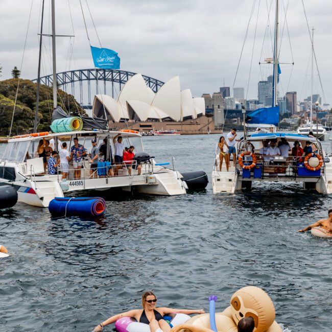 A lively scene on the water with people enjoying themselves on inflatable floats near two docked catamarans. The Sydney Opera House and Harbour Bridge are visible in the background, amidst a cloudy sky. The Yacht Social Club Sydney Boat Hire ensures the water is bustling with activity.