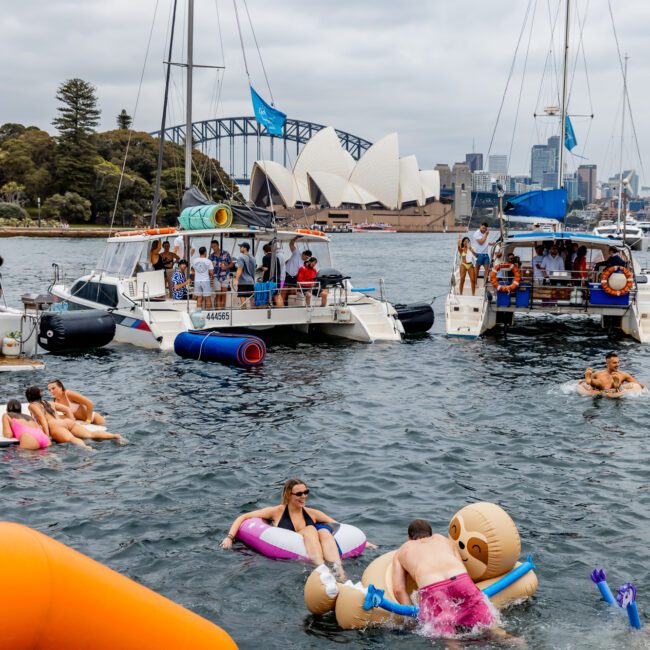 People enjoying a sunny day on a harbor with several boats anchored nearby. Some individuals are swimming or floating on inflatable rafts, while others are relaxing on the boats. The Yacht Social Club Event Boat Charters take center stage with the Sydney Opera House and Sydney Harbour Bridge in the background.