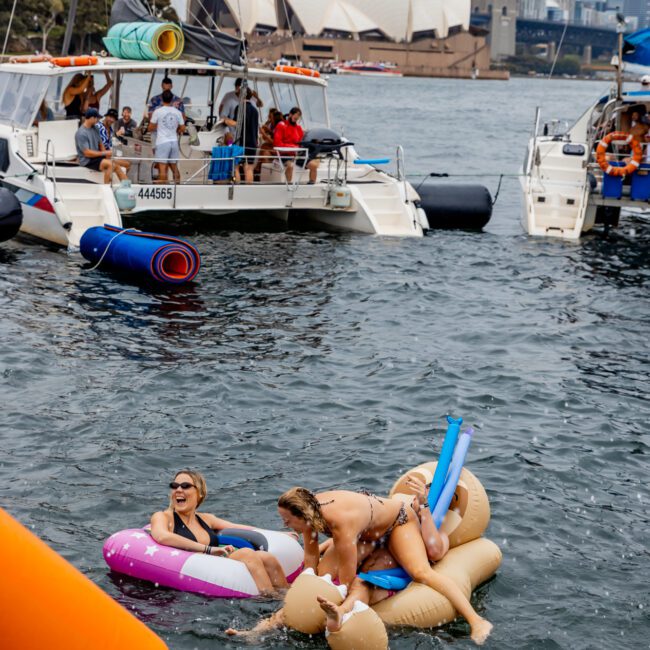 Two women are laughing and playing on inflatable floaties during The Yacht Social Club Boat Party in a scenic harbor. Around them, anchored boats with people watching create a lively atmosphere. In the background, the Sydney Opera House and the Sydney Harbour Bridge can be seen under a cloudy sky.