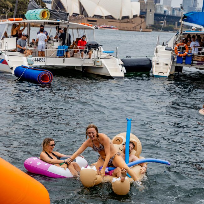 A lively scene of people enjoying a day on the water near the Sydney Opera House and Harbour Bridge. Groups from Boat Parties Sydney The Yacht Social Club are on boats and inflatable floats, smiling and interacting. One group is on large inflatable toys, having fun amidst the bustling activity.