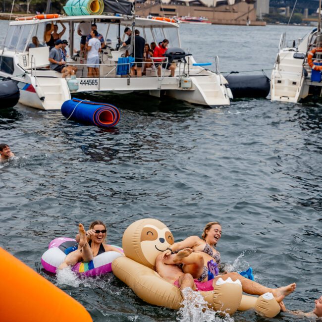 A lively group enjoys a day on the water with the Sydney Opera House and Harbour Bridge in the background. People are playing on large inflatable floaties shaped like a sloth and unicorn. Boats and yachts from Sydney Harbour Boat Hire The Yacht Social Club are nearby, adding to the festive atmosphere.