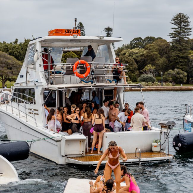 A group of people having a party on a yacht anchored near the shore on a cloudy day. Some individuals are on the upper deck, while others are gathered on the lower deck. In the foreground, three people playfully interact on a floating platform in the water—a perfect scene for The Yacht Social Club Event Boat Charters.