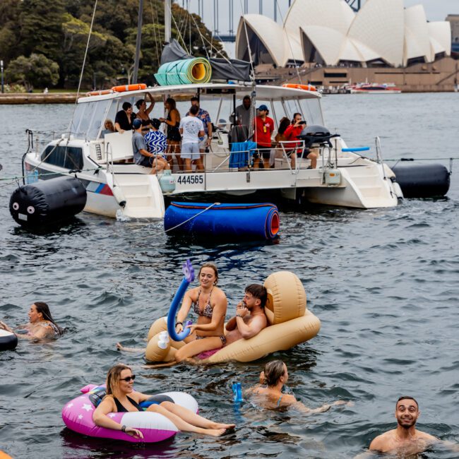 People enjoy a day on the water near a yacht, swimming and relaxing on inflatable floats. In the background, the Sydney Opera House and Harbour Bridge are visible. The yacht has several people on board from The Yacht Social Club, engaging with those in the water.