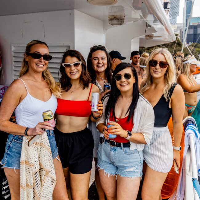 A group of five women stands together on a boat, smiling at the camera. They are dressed in casual summer outfits, holding beverage cans. The background shows other people on the boat and a cityscape partially visible. The mood appears festive and lively, embodying the vibrant atmosphere of Sydney Harbour Boat Hire with The Yacht Social Club Event Boat Charters.