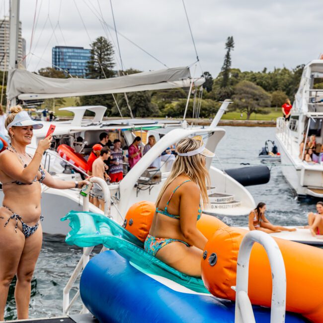 A woman in a bikini slides down an inflatable water slide into the water from a yacht during The Yacht Social Club Sydney Boat Hire event. Another woman nearby takes photos on her phone. Several other boats and people in swimwear are visible in the background, with trees and buildings beyond.