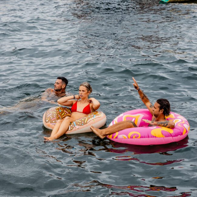 People are floating in the ocean on inflatable pool rings near a boat in Sydney Harbour. One person points upward while another rests on a donut-shaped float. Life rings and lifebuoys are visible on the boat. The Yacht Social Club's buoyant scene is lively and social, perfect for your next boat party in Sydney.