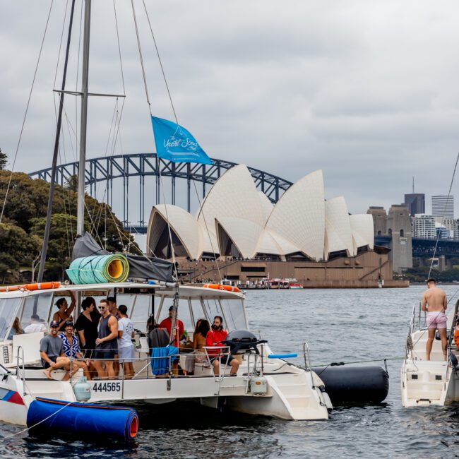 A group of people relaxes on three boats anchored in a bay, with the iconic Sydney Opera House and Sydney Harbour Bridge visible in the background. The sky is overcast, and floating toys are attached to the boats. Enjoying carefree moments, they revel in the experience offered by The Yacht Social Club Sydney Boat Hire.