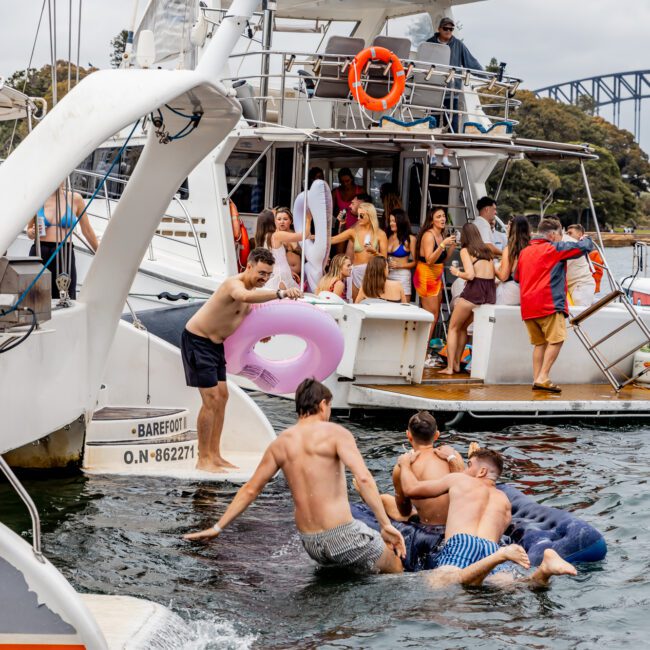 A lively group of people enjoys a yacht party, swimming and relaxing on the boat. Some are on inflatable floats, while others socialize on deck. The scene is set under a cloudy sky with a bridge in the background. The Yacht Social Club Sydney Boat Hire ensures the atmosphere is festive and fun.