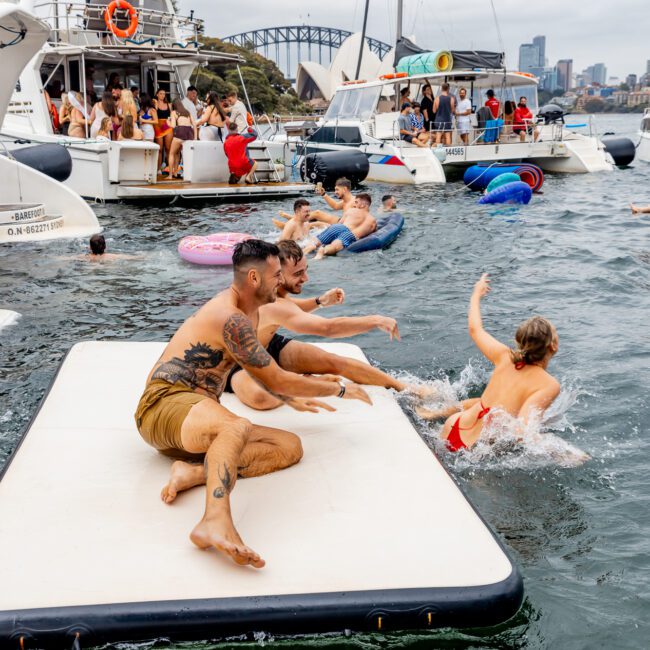 People enjoying a social gathering on a floating platform and surrounding yachts in a harbor. Some are sitting on the platform, with one person playfully splashing water, while others swim or float on inflatables. A cityscape is visible in the background, creating an inviting scene for The Yacht Social Club Sydney Boat Hire.