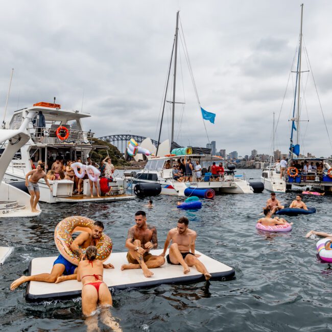 A lively scene of people enjoying The Yacht Social Club Event Boat Charters on the water, featuring several yachts and boats. Some people are on floats and inflatable rings in cheerful colors, while others socialize on a floating platform and boats. The sky is cloudy, with a cityscape in the background.