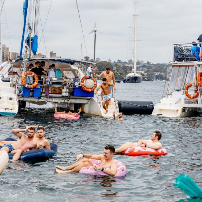 A lively scene of people enjoying a day on the water. Several individuals are swimming and floating on colorful inflatables near two anchored boats from The Yacht Social Club. A cloudy sky and urban landscape are visible in the background. The mood is joyful and relaxed.