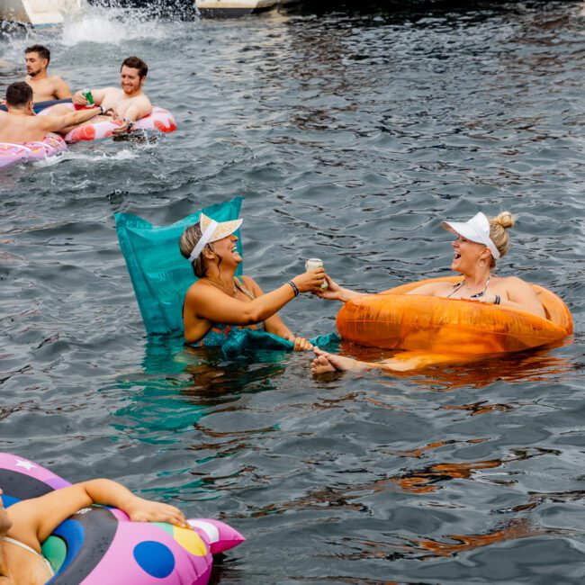 Two women clink drinks while relaxing on inflatable pool floats in a body of water, surrounded by others also on floats. In the background, a boat with "The Yacht Social Club" sign and people on board is visible. The atmosphere is lively and festive.