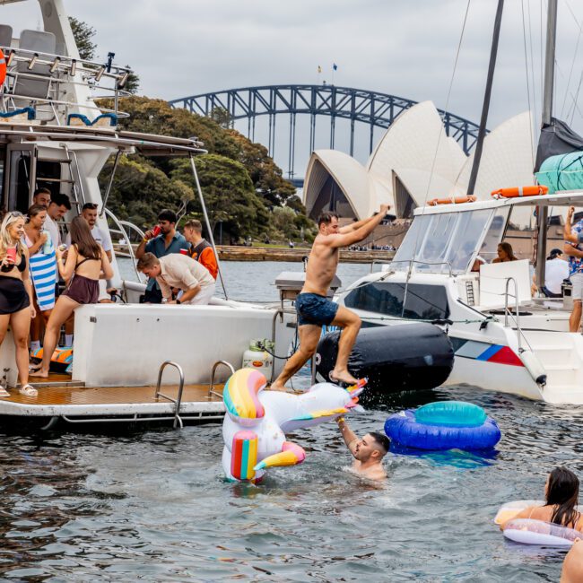 A lively scene on the water near Sydney Opera House and Harbour Bridge: people gather on boats, with one person jumping onto a unicorn float. Others enjoy various inflatables and boat decks, part of The Yacht Social Club by Sydney Boat Hire. The sky is cloudy.