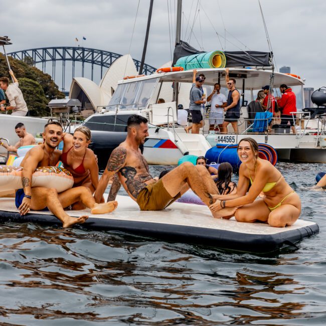 A group of people in swimwear relax on an inflatable float in the water, smiling and enjoying themselves. Behind them, a luxury yacht from Sydney Harbour Boat Hire The Yacht Social Club can be seen with more people onboard near a bridge. The sky is overcast, but everyone appears to be having a good time.