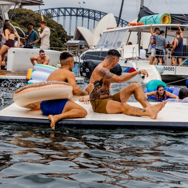 Two men sit on a floating platform in the water near several boats, with the Sydney Opera House and Harbour Bridge visible in the background. People in swimsuits enjoy the sunny day and festive atmosphere, courtesy of The Yacht Social Club Sydney Boat Hire.