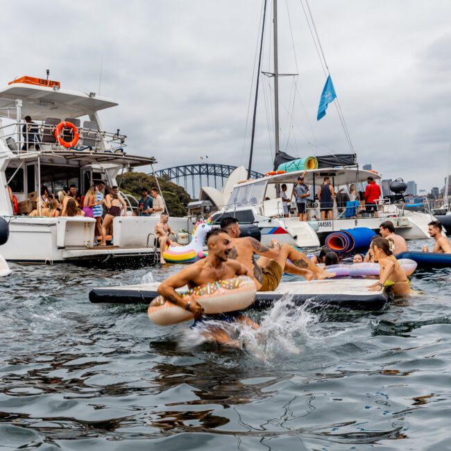 A lively scene on the water with people enjoying a Yacht Social Club event. Several individuals are on inflatable rafts and floats, splashing and having fun. Yachts are docked in the background, with a bridge visible under a cloudy sky. A blue flag is flying in the distance.